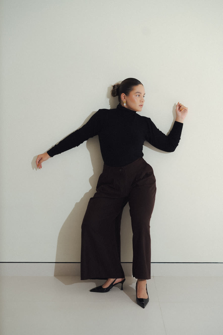 Mafer González de cuerpo entero, con suéter negro y pantalón cacao, apoyada en una pared clara